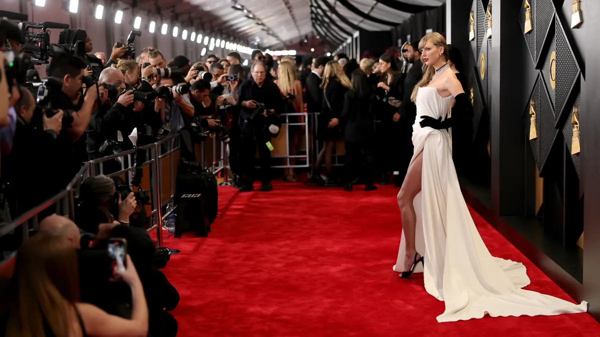 Taylor Swift stands on a red carpet in a long white strapless gown while being photographed by multiple people.