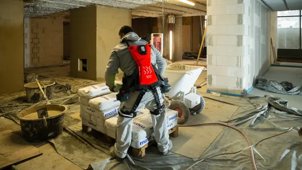 A person in gray work clothes uses a red exoskeleton to pick up and put down heavy bags on a construction site.