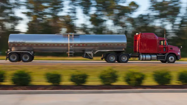 A tanker attached to a red tractor driving on a highway with blurred trees in the background.