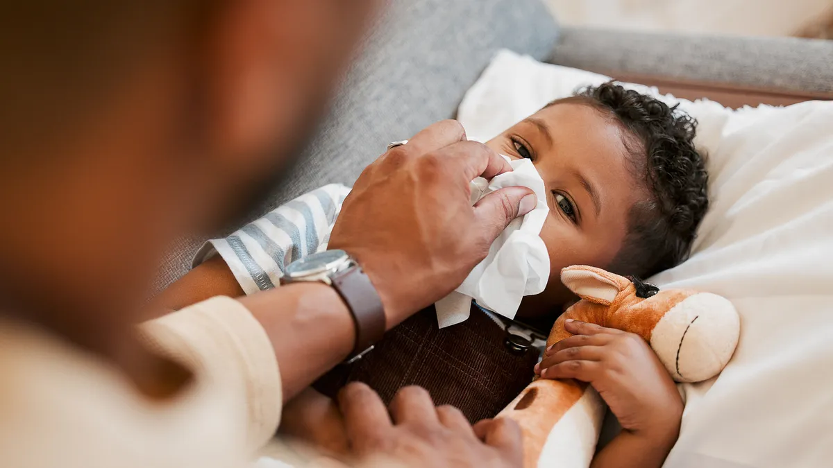 Sick, ill and unwell little child suffering from cold, flu or COVID-19, and lying on the sofa at home while blowing his nose with dad