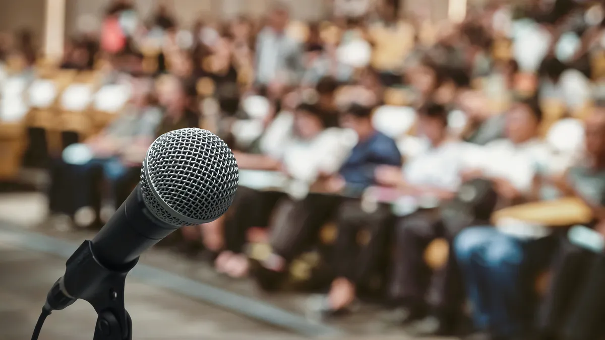 A standing microphone is seen in the foreground. In a blurred background are people seated in rows of chairs