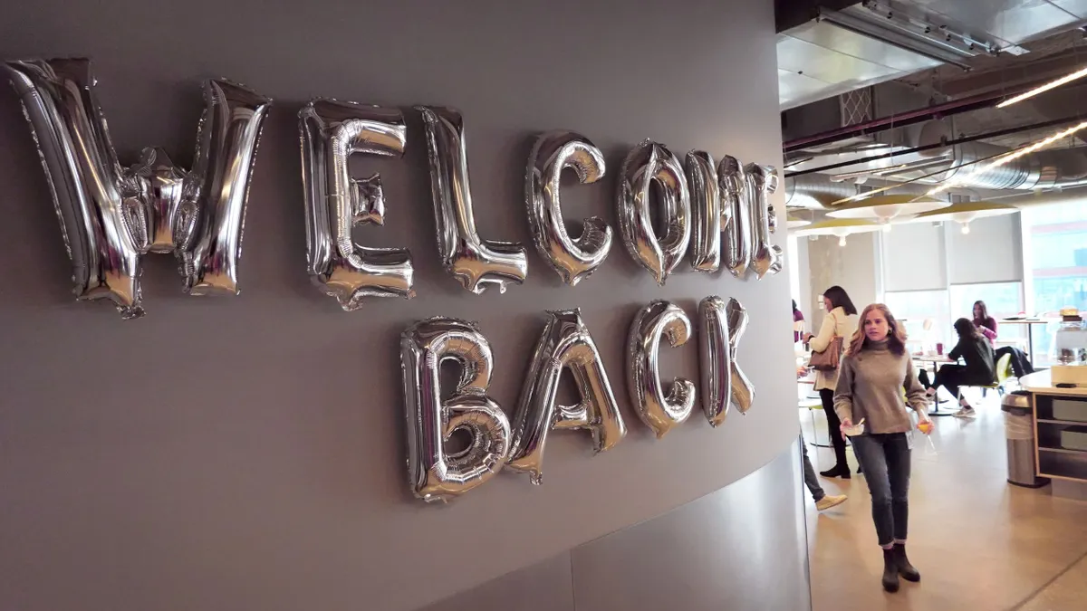 People walk through a Google office cafeteria, past a sign welcoming them back.