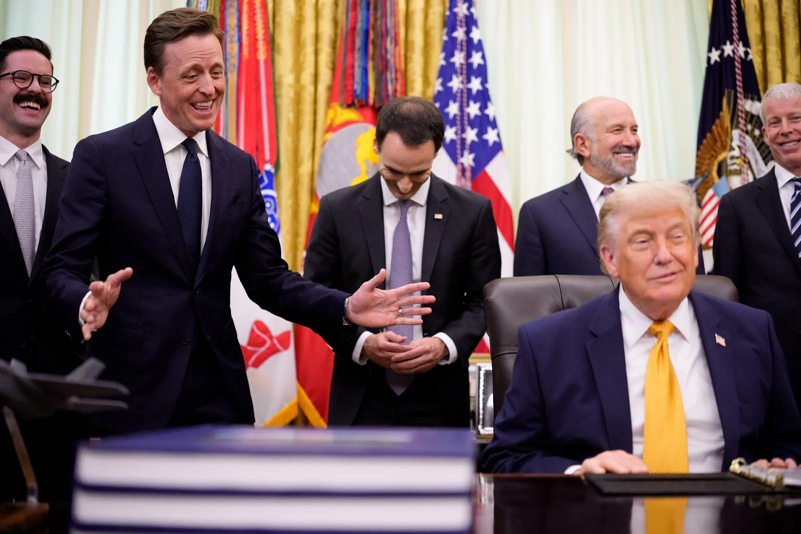 A man gestures with his hands while standing behind another man who sits at a desk. Behind the two men, other men stand in a line in front of curtained windows.