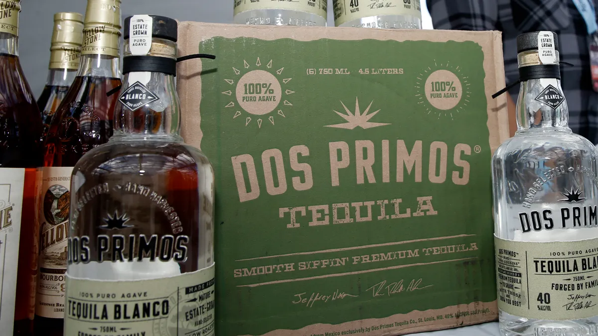 An assortment of bottles and a box on a table while a person looks on. The box reads, "Dos Primos Tequila."