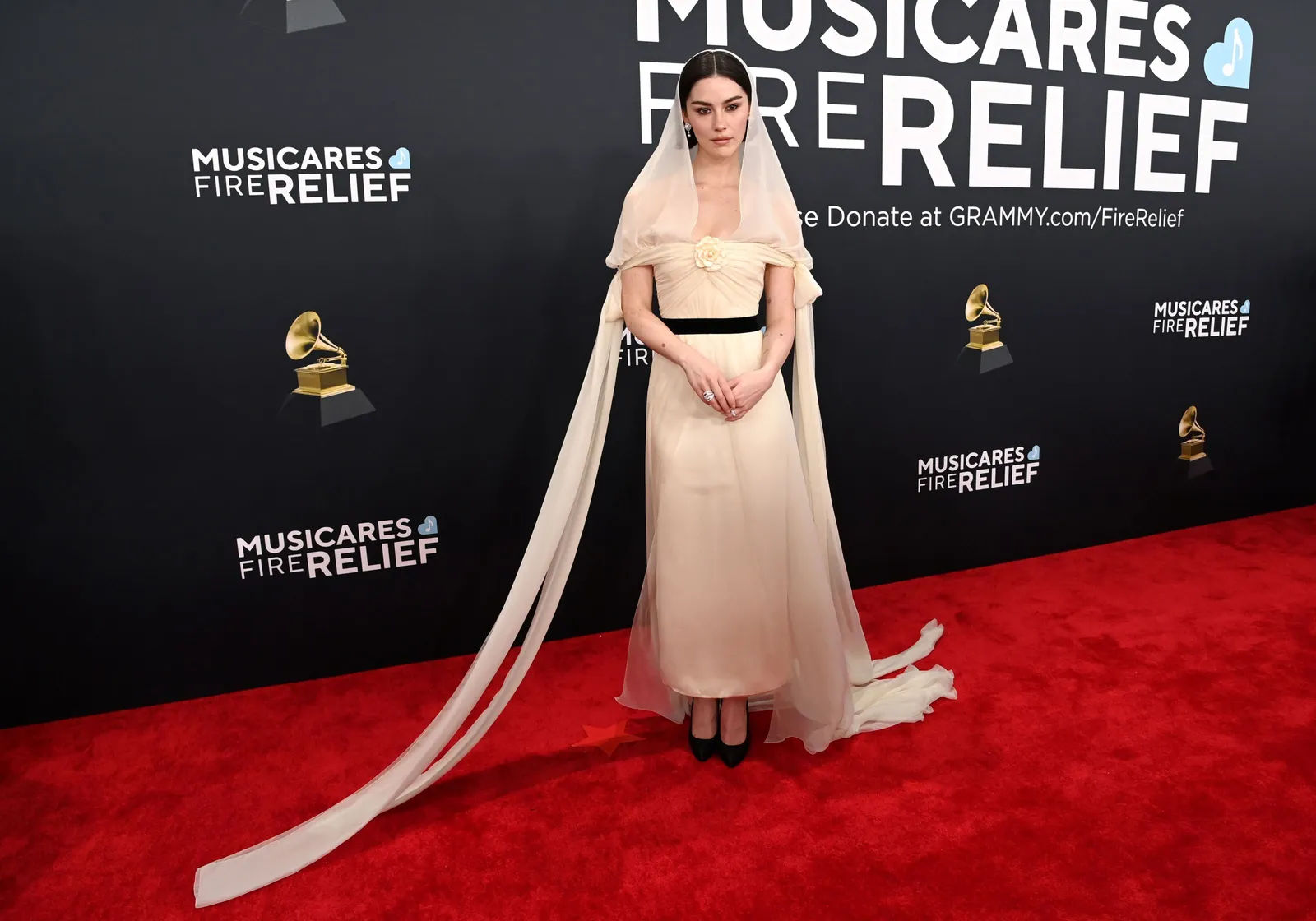 A person in a white gown and headscarf stands on a red carpet.