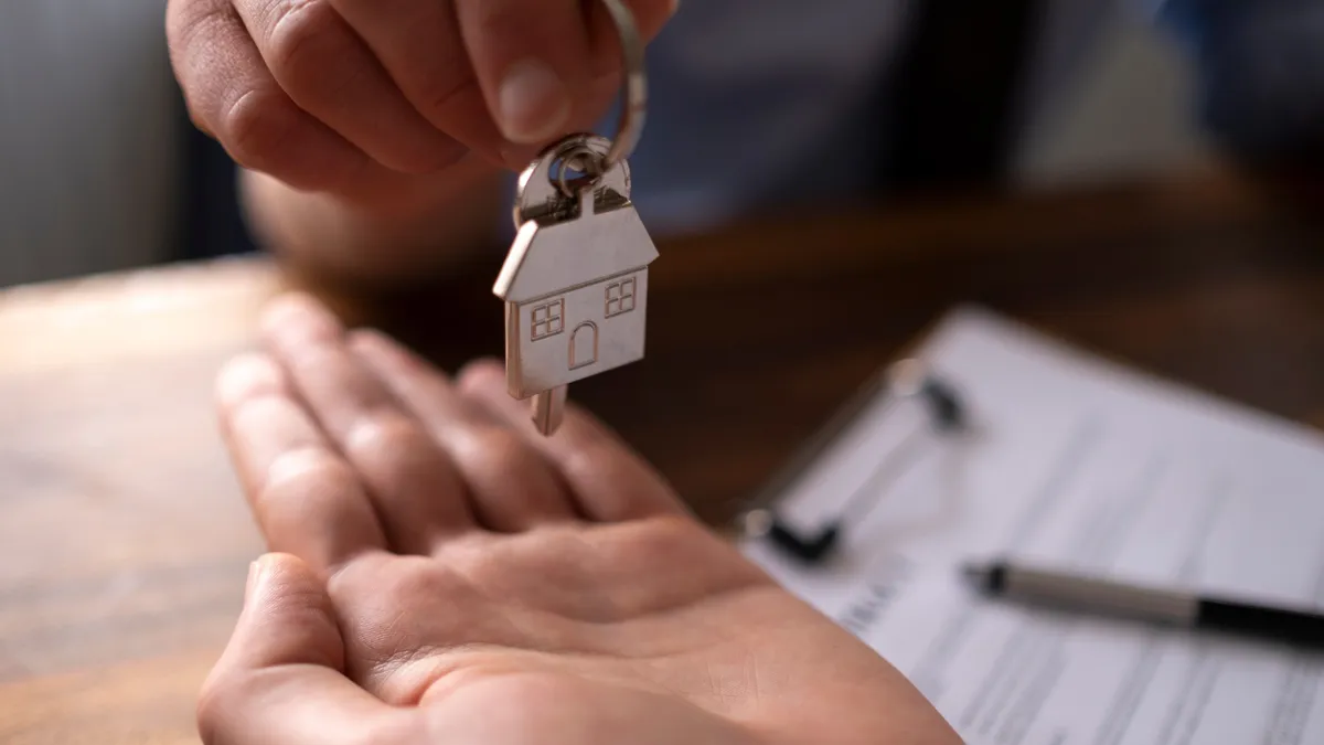 one hand placing a house key in another hand on top of a desk with paperwork