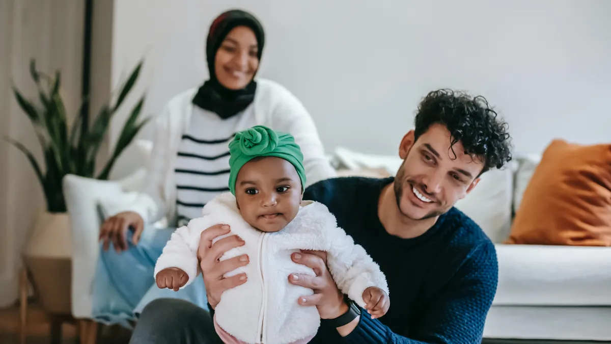 A brown dad plays with their baby, who is wearing a headwrap. In the background, his partner who is wearing a hijab, smiles.