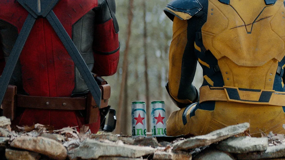 The back of Deadpool, in red, and Wolverine, in yellow, sitting in a forest setting with two silver cans of Heineken Silver between them.