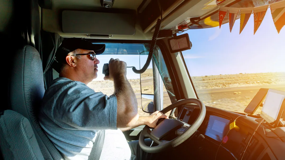 A truck driver holds a radio and steering wheel with different devices connected on the vehicle's dashboard.