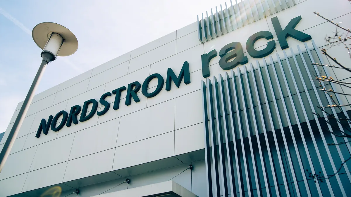 A street lamp tilts right as the white storefront of a "Nordstrom Rack" location tilts left, as sun streams in a light blue sky. Vertical bars decorate the building above and below the word "Rack."