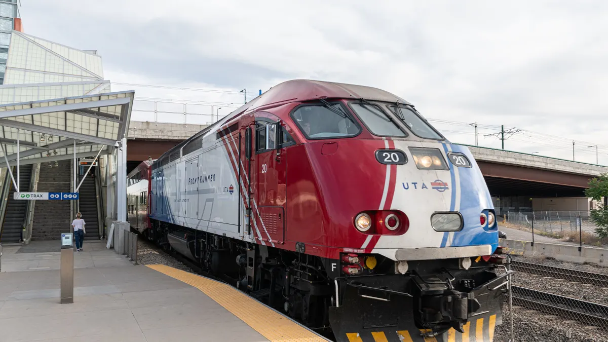 A red, white and blue locomotive and train at a station with a person walking toward an escalator.