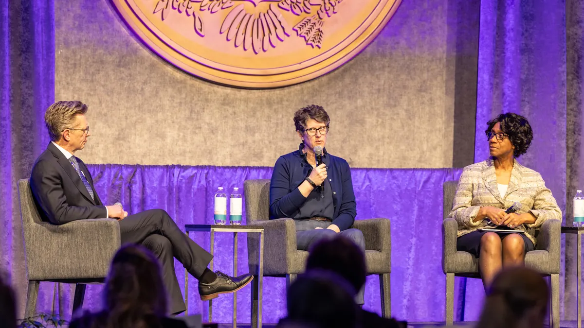 Three people are sitting in chairs on a stage. One person has a microphone