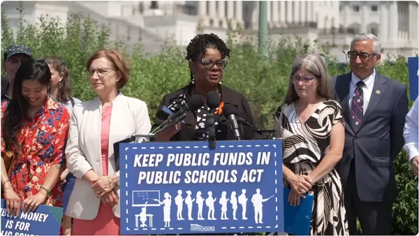 A person stands at a podium outside the Capitol building in Washington, D.C. Other people are standing beside the person. A sign on the podium says "Keep Public Funds in Public Schools Act"