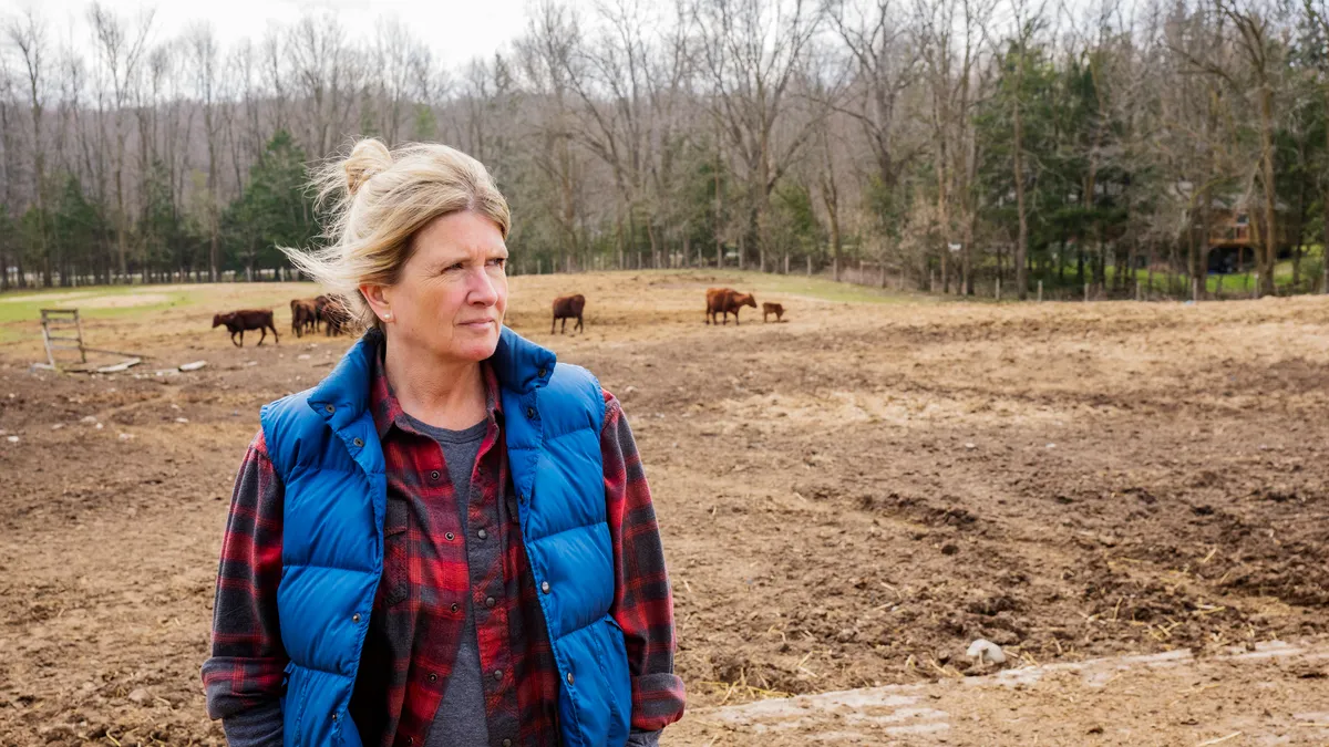 A femme-presenting person in a flannel shirt and puffer vests looks off to the side in a rural setting; there is livestock behind them and trees