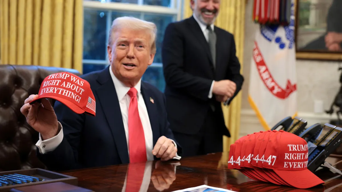 Donald Trump holds up a hat as he signs an executive order while sitting at his desk in the Oval Office