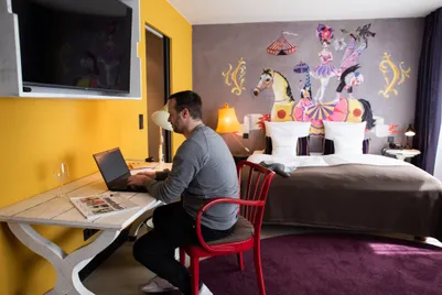 A man sits with a laptop at a white desk in his hotel room with a bed in the background