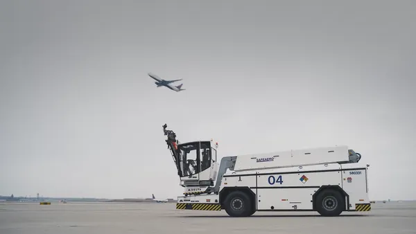 A deicer vehicle parked  on an empty airport runway and an airplane flying overhead.