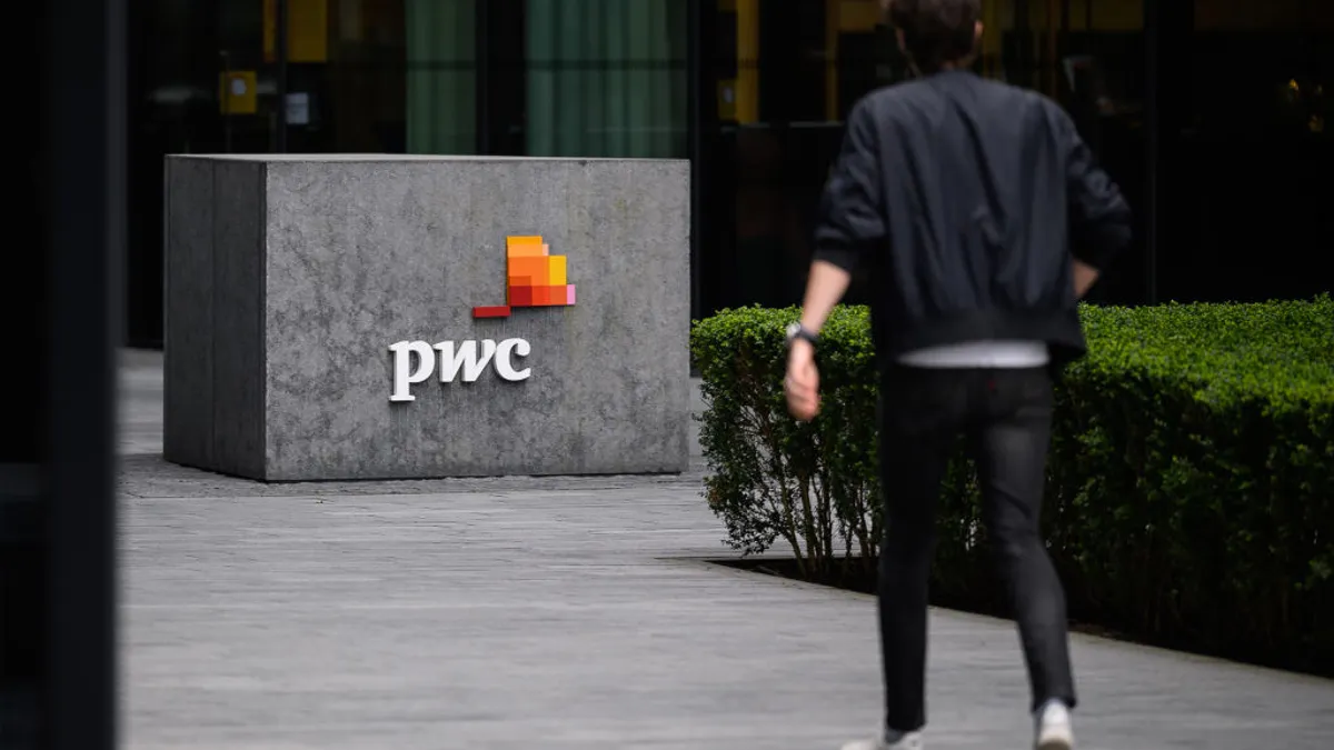 A person with dark clothing walks through a corporate campus, show out of focus. The PwC logo in a company sign is shown in the background.