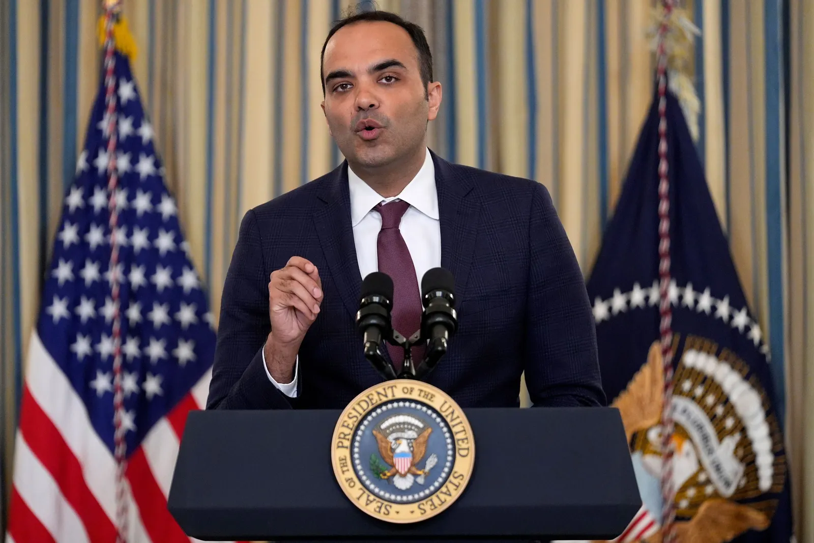 Former Consumer Financial Protection Bureau Director Rohit Chopra stands at podium, with one hand raised and flags in background.