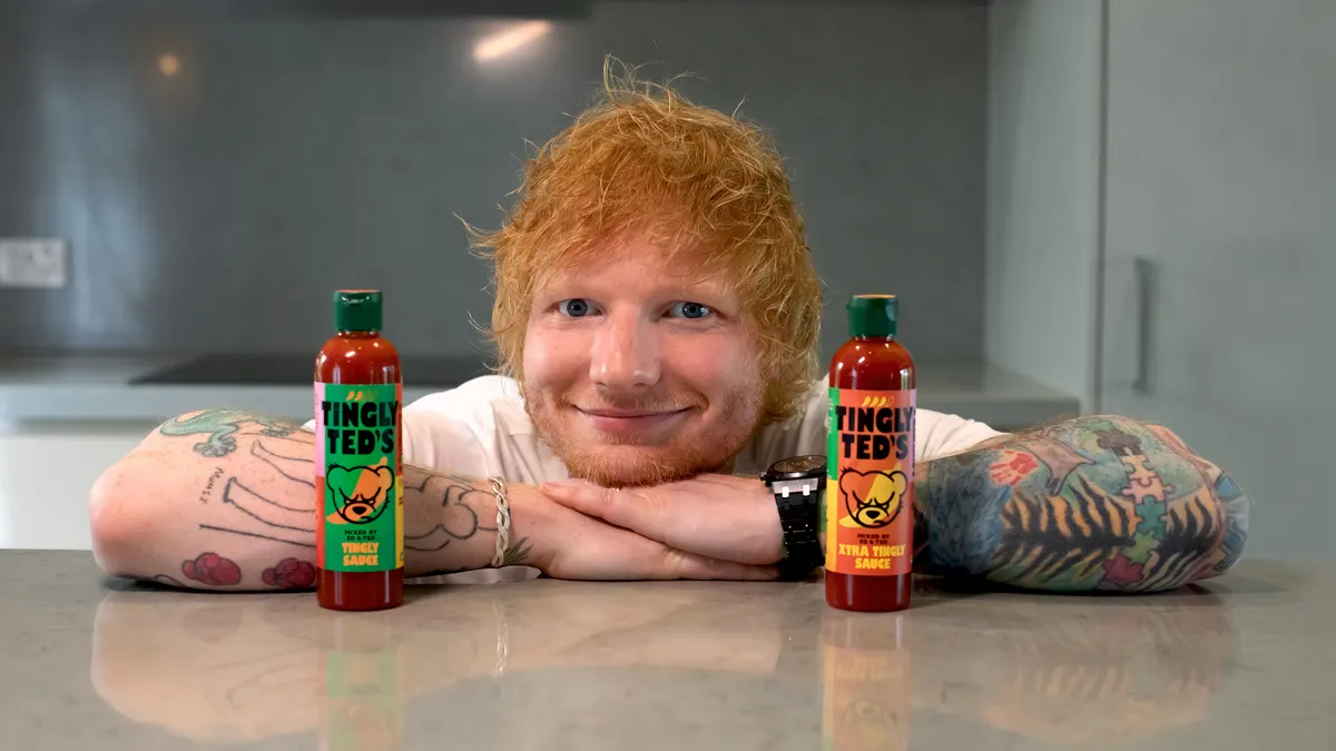 Ed Sheeran leans on a shiny gray countertop with a bottle of Tingly Ted's hot sauce on either side of him.
