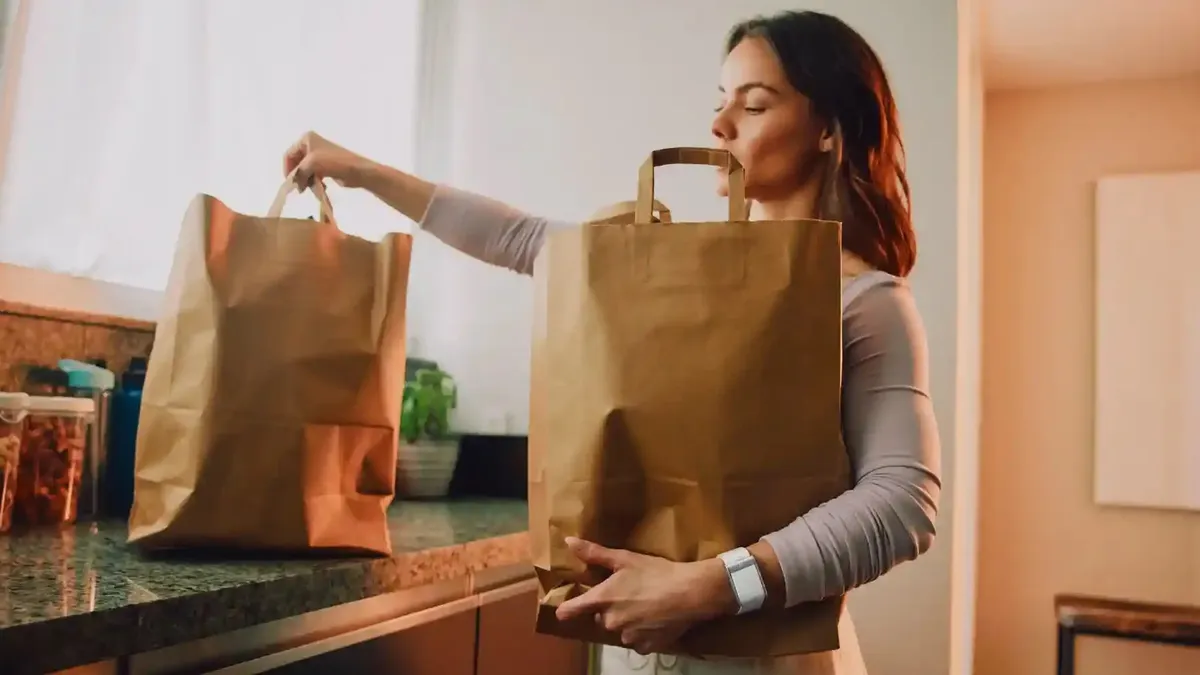 A person sets a brown, paper bag on a counter and holds another bag. They are wearing a white wristband.
