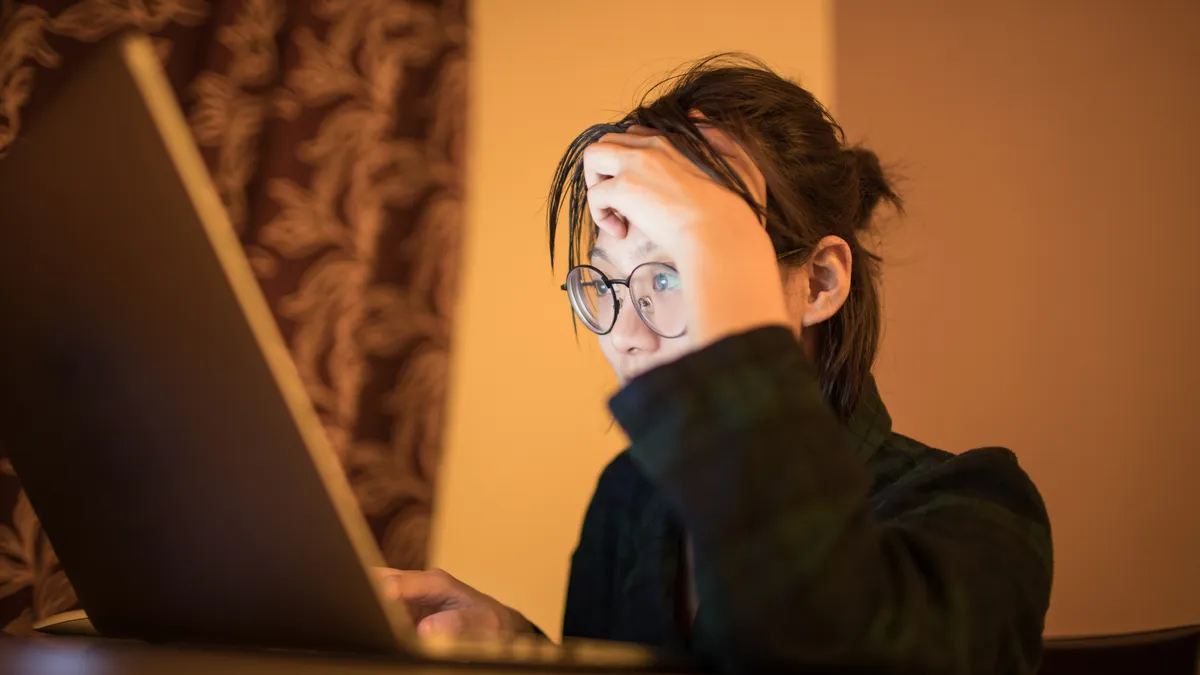 A student, wearing glasses, sits at a desk with an open laptop and hand over their head.