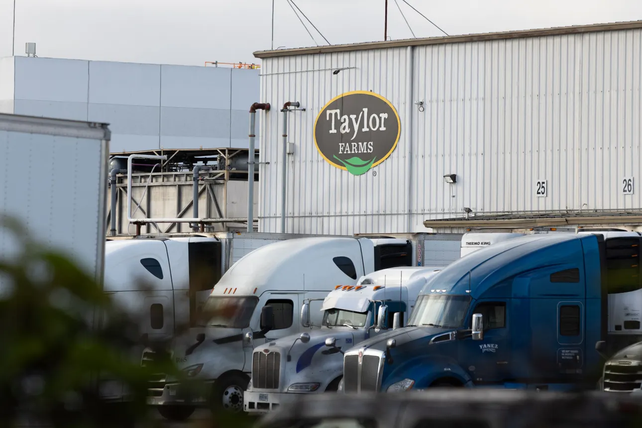 Trucks sit parked outside a logistics facility. The building has the "Taylor Farms" logo printed on its side.