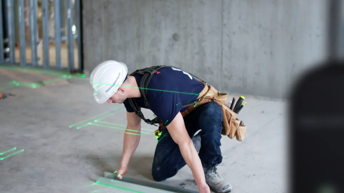 A person with a white hardhat kneels on a floor while a machine projects green light onto the ground.