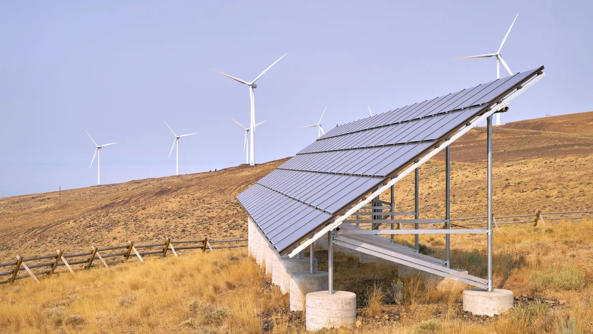 A large solar panel adheres to a cement bass on a hill surrounded by wind turbines. 