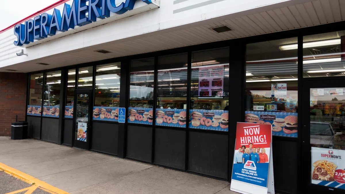 Exterior of a SuperAmerica convenience store with a red and blue 'NOW HIRING!' sign and advertisements in the windows.