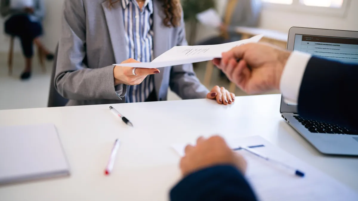 A person hands a resume across a desk to another person in formal clothes.