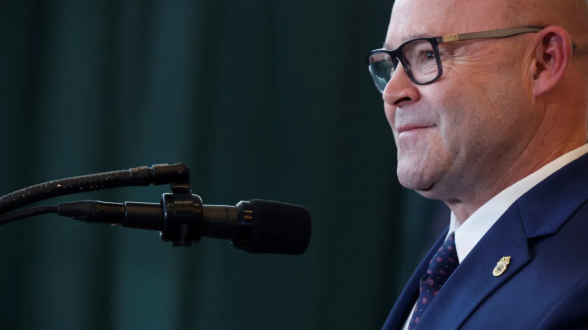 A bald man with glasses stands in front of a microphone.