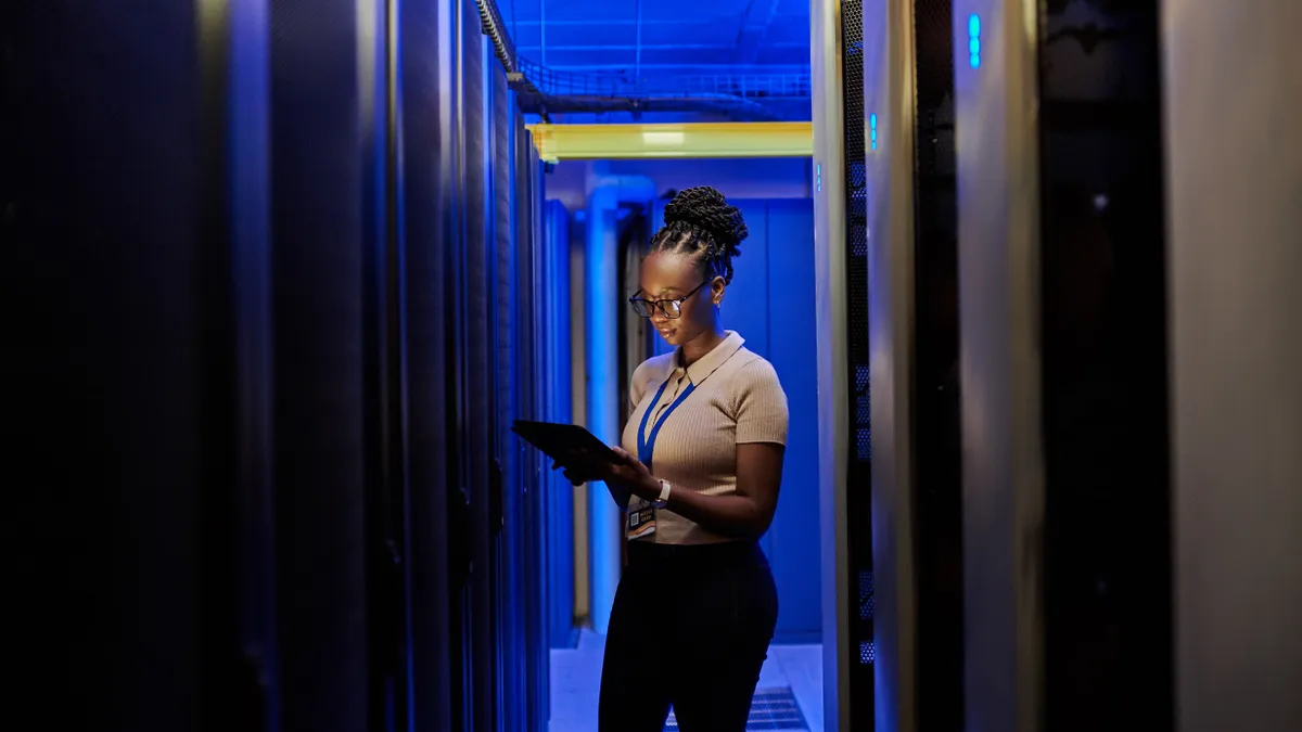 An employee checks a tablet computer while standing between two rows of computer servers.