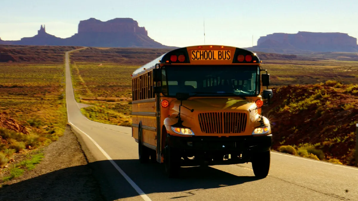 A yellow school bus is driving on a desolate road. Untouched landscape is seen on both sides of the road.