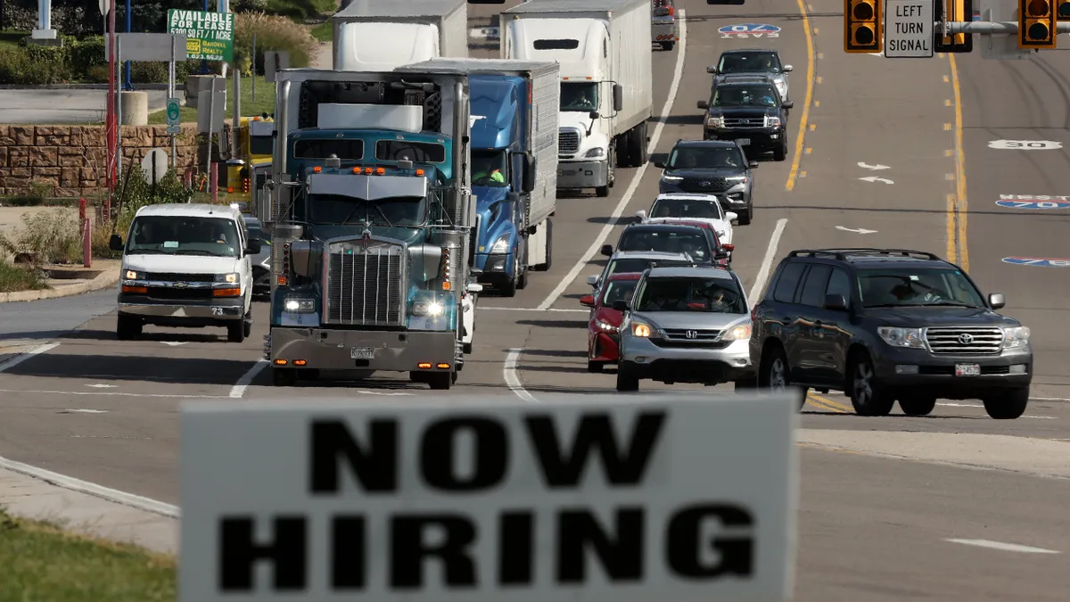 Trucks drive on a road past a "NOW HIRING" sign.