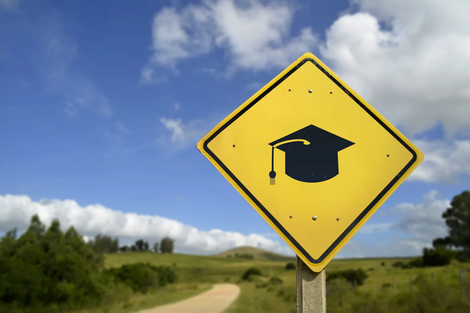 A road sign with a graduation cap icon stands in a rural, grassy landscape.