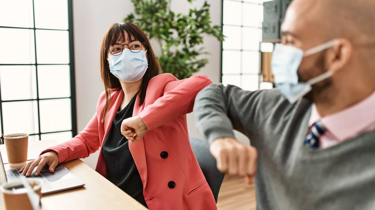 Two people wearing face masks elbow bump each other while sitting at a wooden desk.