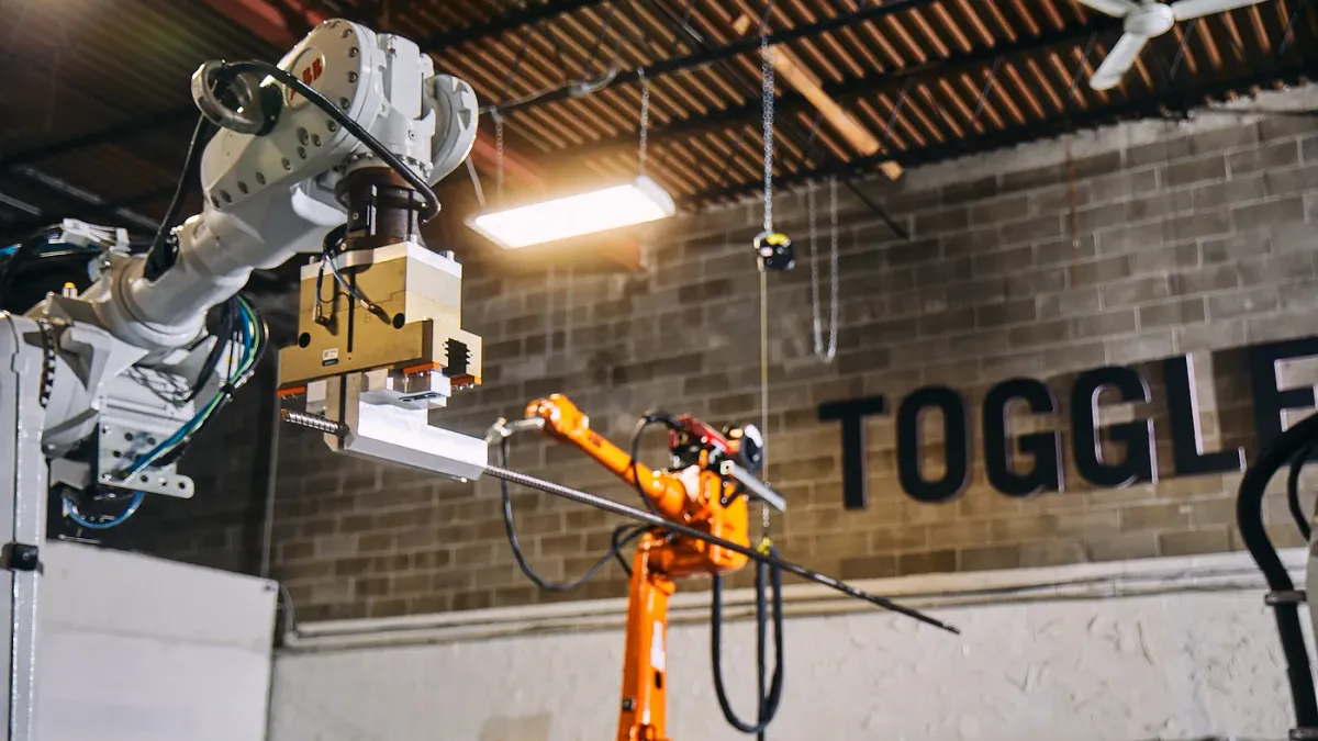 Robots put together a rebar in an industrial warehouse with a brick background. The company's name, "Toggle," appears in the background on the wall in black letters.