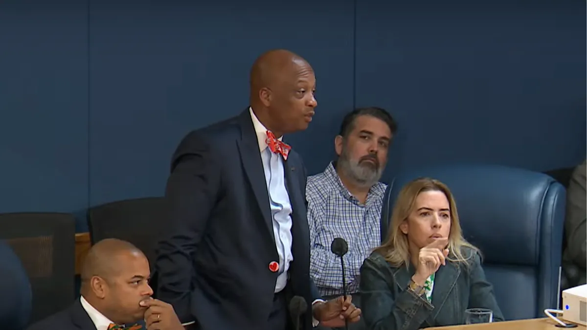 County Board Chair Oliver Gilbert III stands in front of a mic with two other commissioners on a dais seated on either side of him.