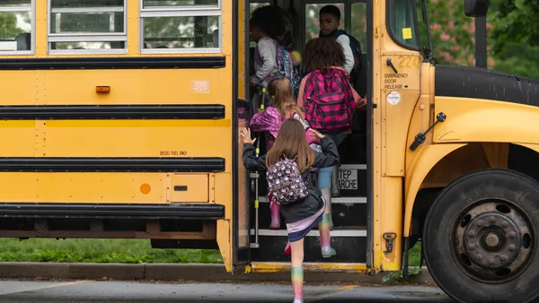 A yellow school bus is seen from the side with an open door and a handful of children walking up the steps into the bus.