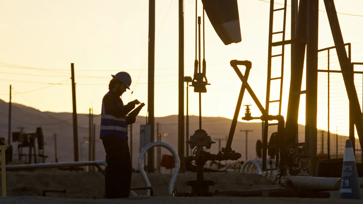 A person wearing a uniform and hard hat looks down at a device in their hands. Behind the person are pipes and industrial equipment silhouetted against the sunset.