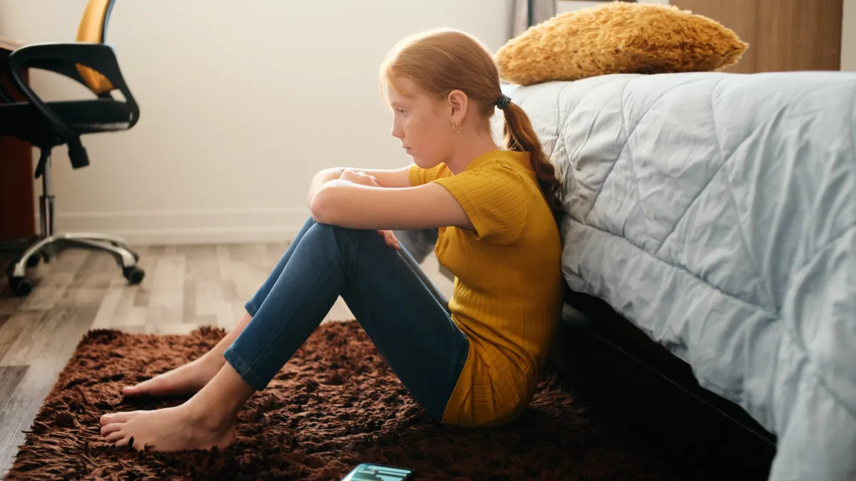 A teenage girl sits at the side of her bed on the floor with a cellphone next to her