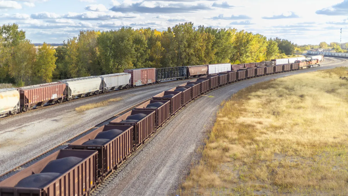 Freight train transporting coal and commodity with backdrop of green trees.