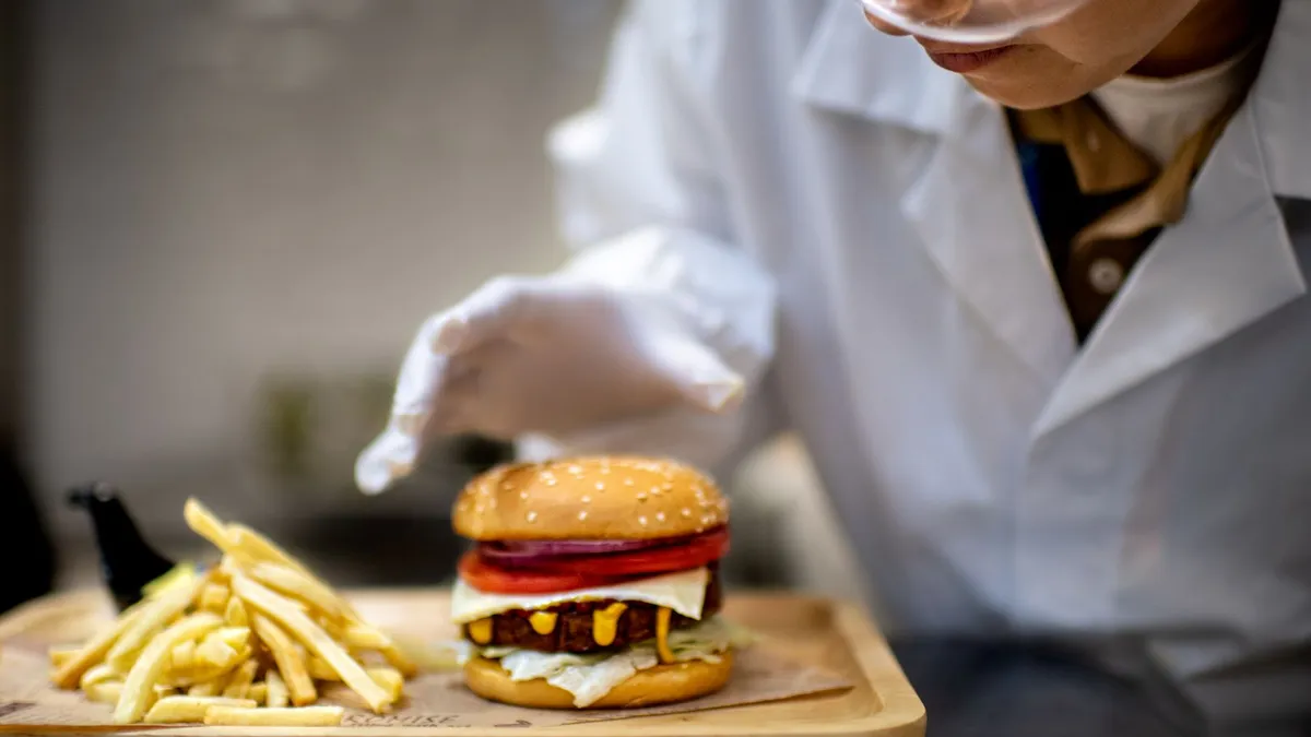 A person wearing a white lab coat and goggles focuses on a plant-based hamburger and fries.