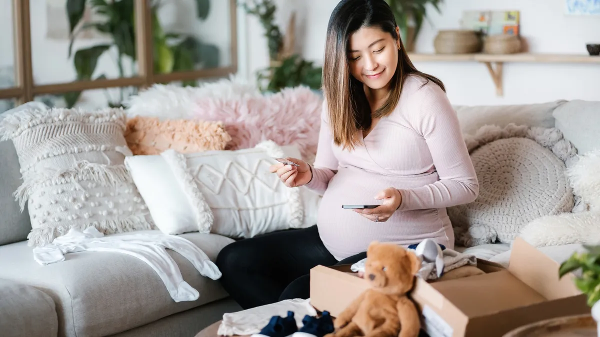 A pregnant person sits smiling on a couch with a set of toys on a table.