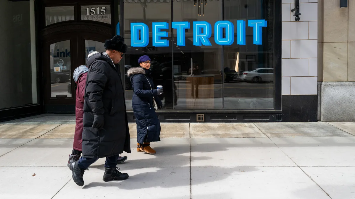 Three people walk on a sidewalk in front of a window that says "Detroit."