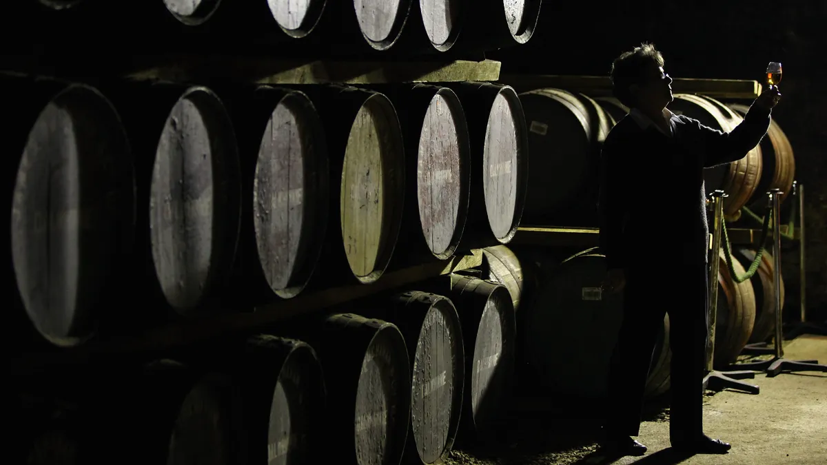 A person holds up a glass of aged spirits near a wall of barrels.