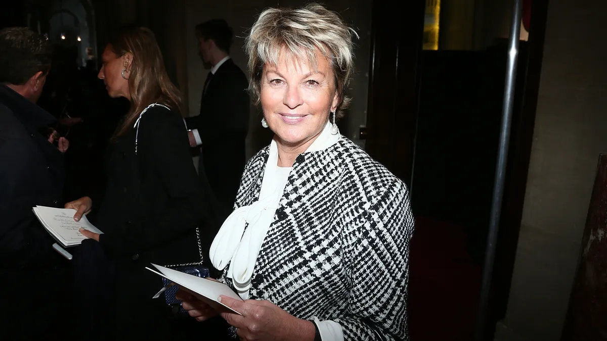 Baronne Myriam Ullens De Schooten wears a white tie-neck blouse and a black and white checked jacket, with short blonde hair, and smiles at the camera