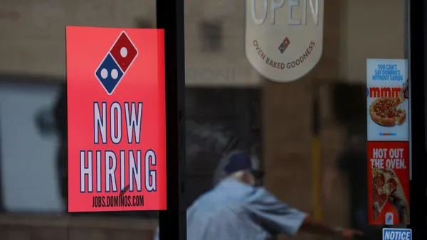 A photograph of a glass window to a Domino's restaurant with a red sign that says now hiring.