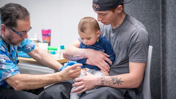 A young child gets a shot from a medical professional while another adult holds the child in their lap.